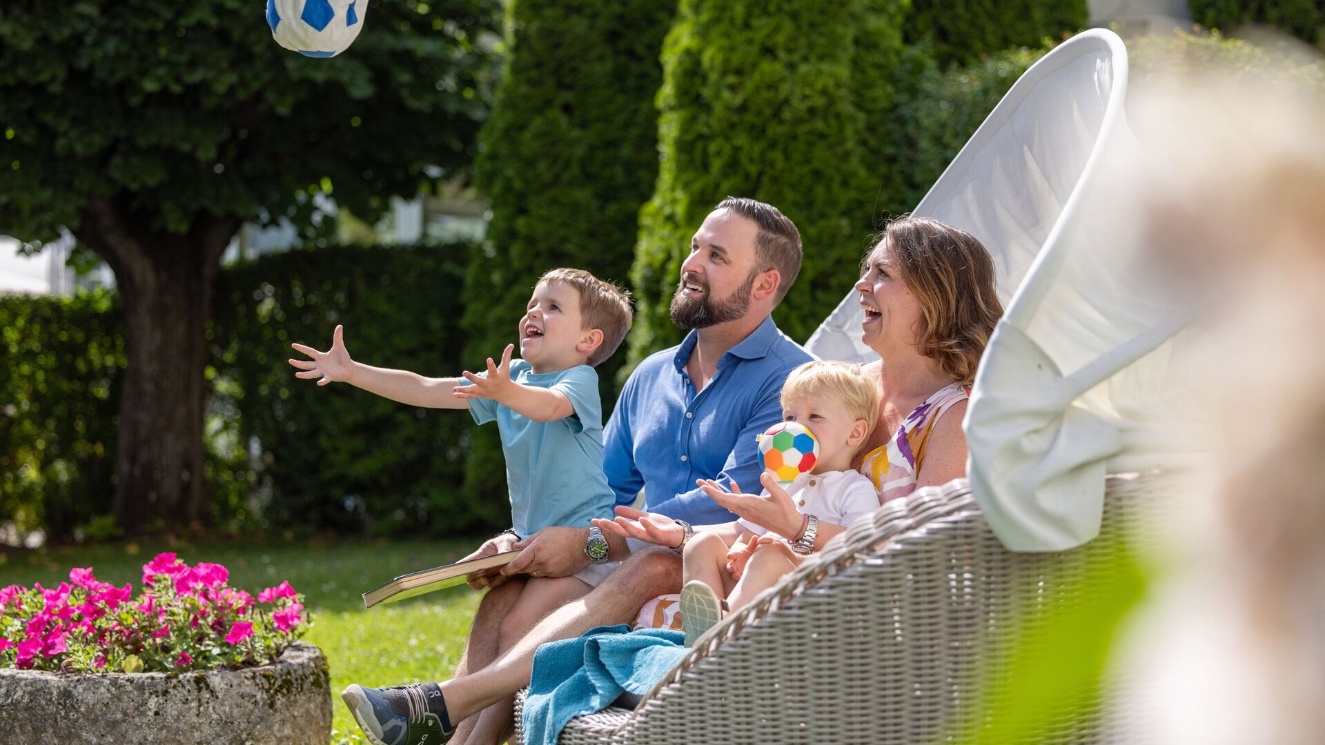 Eine Familie sitzt entspannt im Gartenbereich, während ein Kind nach einem Ball greift.