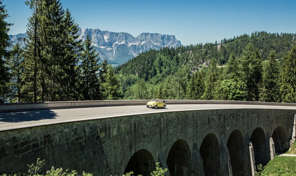 Ein gelber Oldtimer fährt über eine Brücke auf einer Panoramastraße vor bewaldeten Bergen.