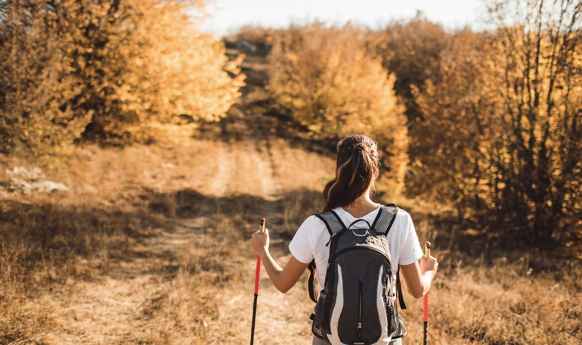 Eine Wanderin mit Rucksack geht auf einem herbstlichen Weg zwischen goldenen Bäumen in einer Berglandschaft.