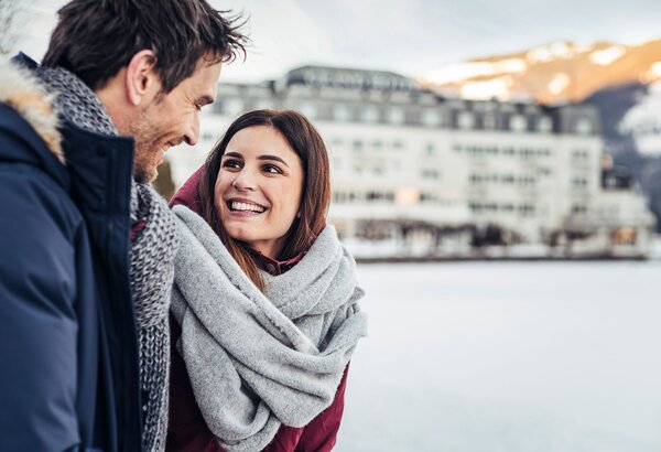 A smiling couple stands by the frozen Zeller See with snowy mountains in the background.