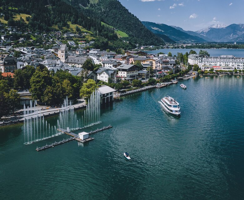 Blick über den Zeller See auf die Uferpromenade mit Springbrunnen, Booten und den Bergen von Zell am See.
