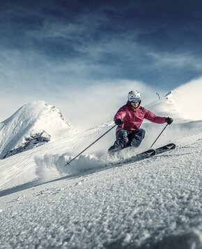 Ein Skifahrer in pinker Jacke fährt eine präparierte Schneepiste vor verschneiten Bergen hinab.