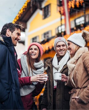 Vier Freunde stehen draußen im Winter und halten Becher in der Hand, im Hintergrund sind warm beleuchtete Häuser zu sehen.