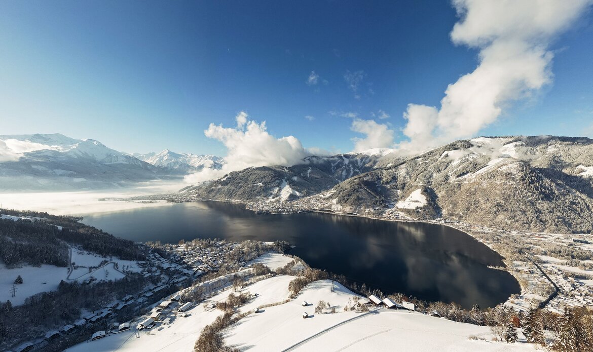 View over the snow-covered Lake Zell with mountains and clouds beneath a blue sky.