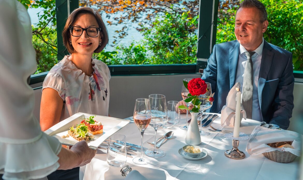 Two guests are seated at a set table in a hotel restaurant while a waiter serves food.