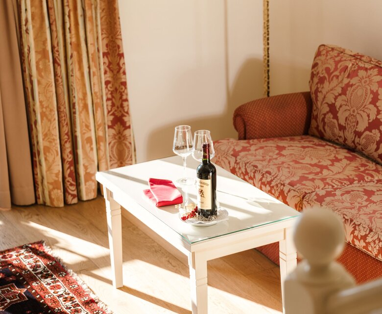 Ein Wohnbereich einer Suite mit Sofa, weißem Tisch, Weinflasche und Blick auf den Balkon im GRAND HOTEL Zell am See.