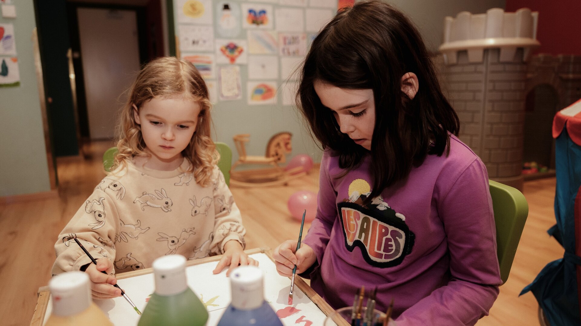 Two children sit at a table and paint together in a play area.