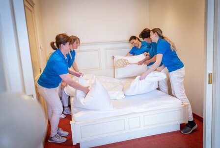 Hotelteam beim Bettenmachen im GRAND HOTEL Zell am See Vier Mitarbeitende beziehen gemeinsam ein Hotelbett in einem hellen Gästezimmer.