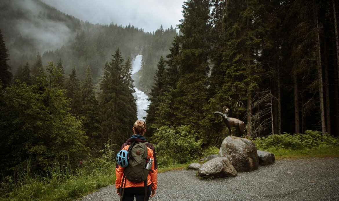 A hiker stands on a forest path and looks toward a steep waterfall in Salzburger Land.