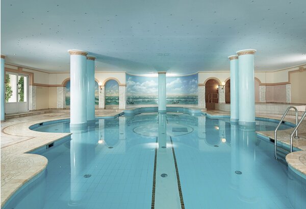 Ein heller Innenpool mit Säulen und Blick auf eine gemalte See- und Berglandschaft im Classic Spa des GRAND HOTEL Zell am See.