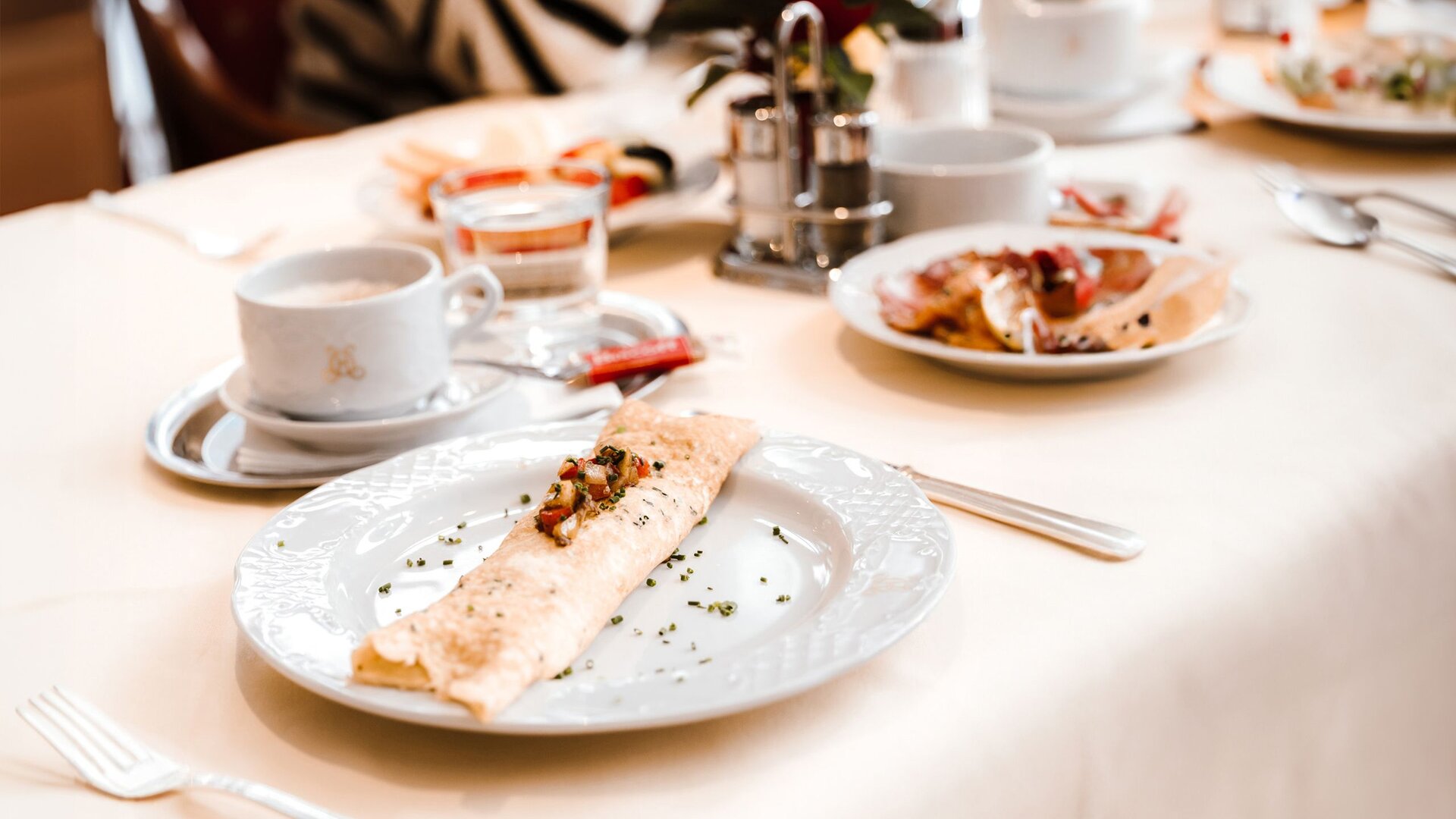 Auf dem Tisch stehen ein gefüllter Crêpe, eine Kaffeetasse, ein Glas Saft und weiteres Frühstücksgeschirr.