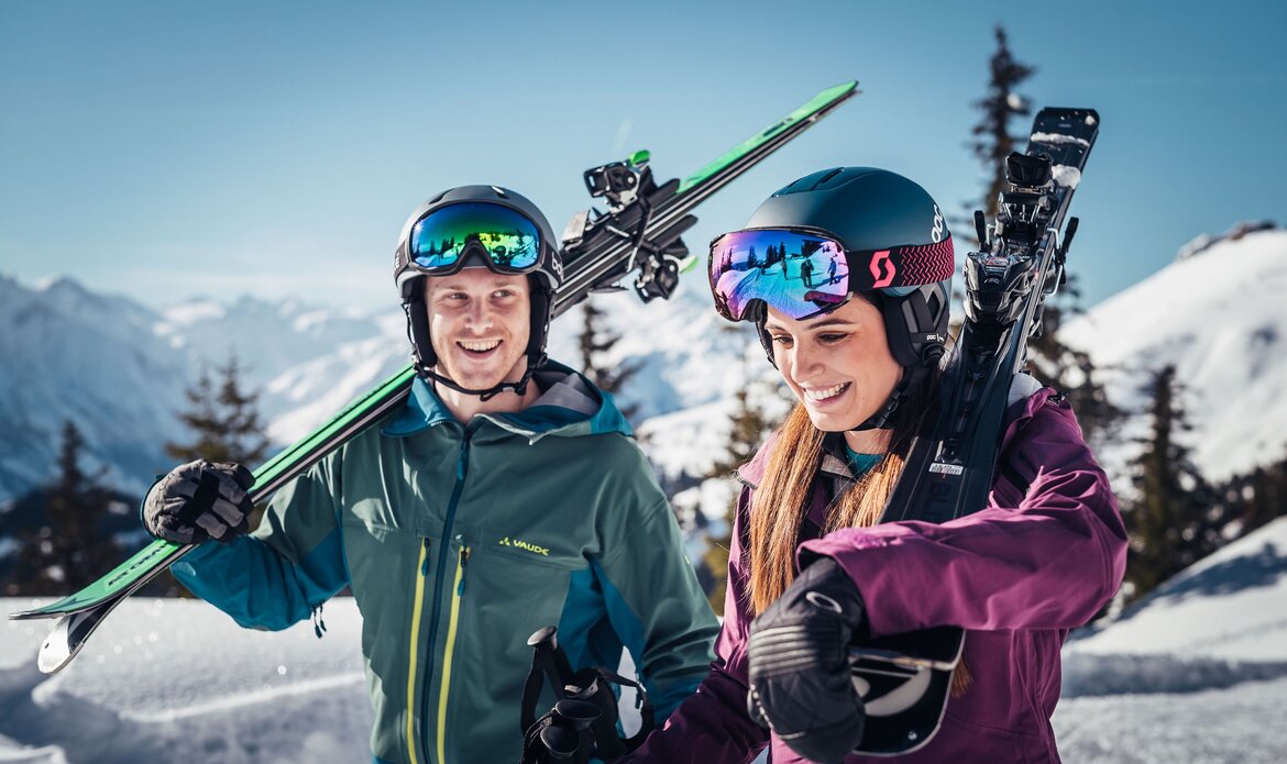 Two skiers stand smiling in a snowy mountain landscape, carrying skis on their shoulders.