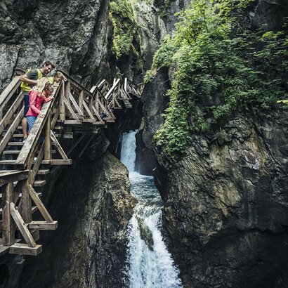 A family stands on a wooden walkway in a narrow gorge beside a small waterfall.