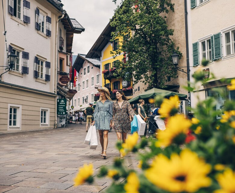 Zwei Frauen spazieren durch eine Altstadtgasse in Zell am See, im Vordergrund sind gelbe Blumen unscharf zu sehen.