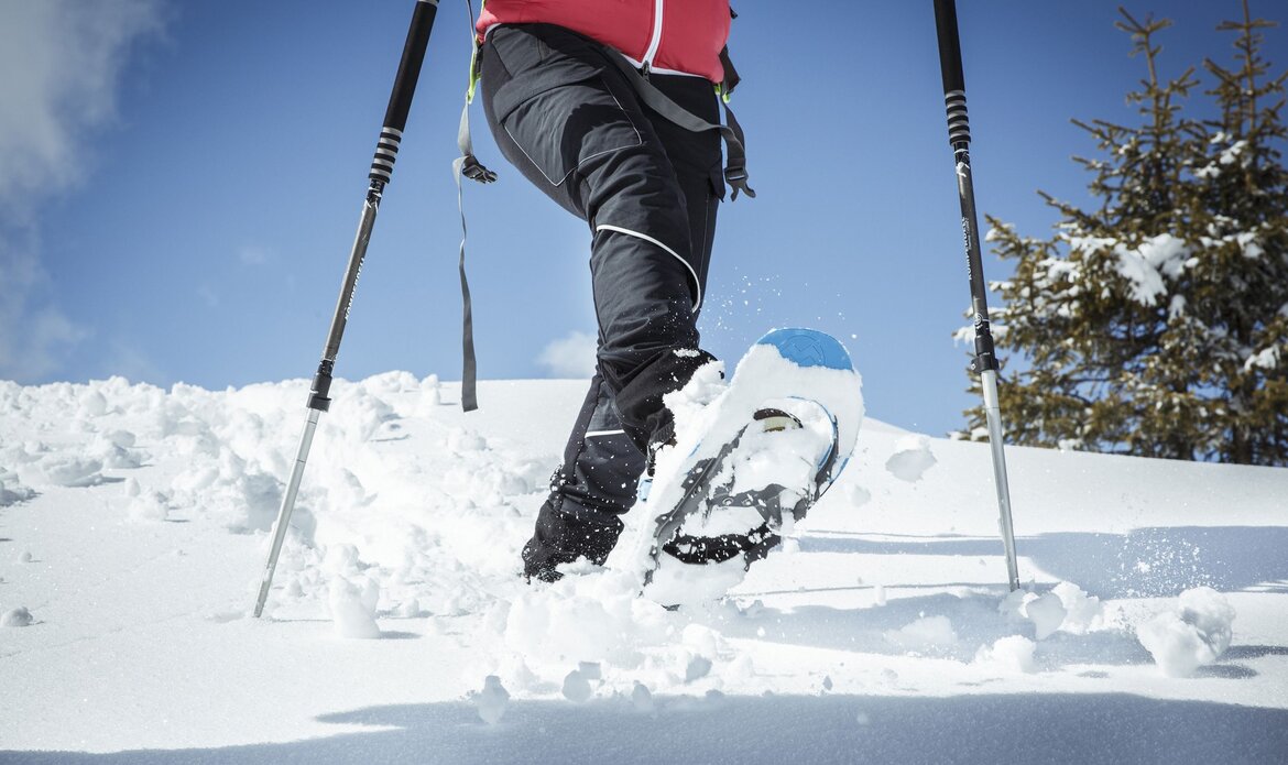 A person walks through deep snow with snowshoes under a clear blue sky and a snowy mountain backdrop.