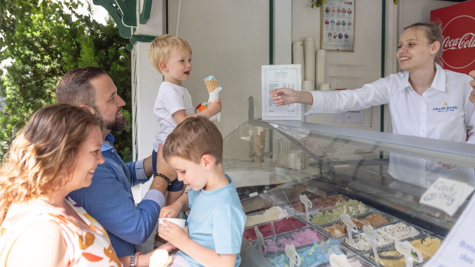 Eine Familie mit zwei Kindern steht an einer Eistheke, während eine Mitarbeiterin eine Portion überreicht.