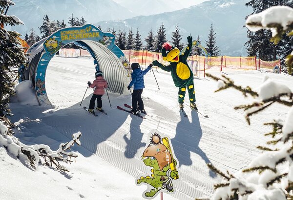 Zwei Kinder und eine Figur im Kostüm stehen an einem Kinderskigebiet im Schnee vor verschneiten Bergen.