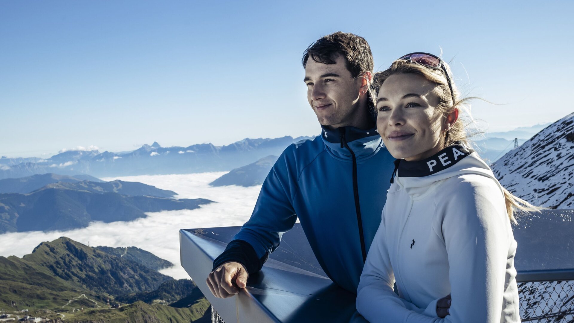 A couple stands on a viewing platform overlooking the mountains and a sea of clouds in Salzburger Land.