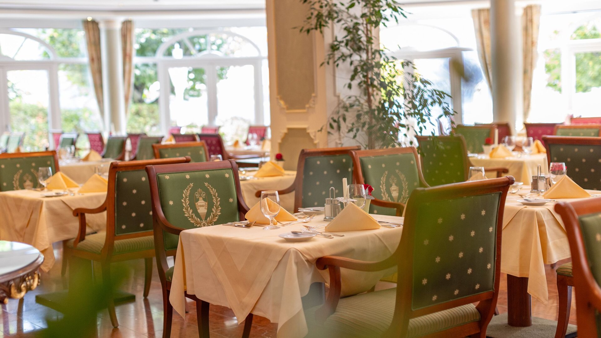 Ein elegant gedeckter Speisesaal mit grün gepolsterten Stühlen, eingedeckten Tischen und großen Fenstern.