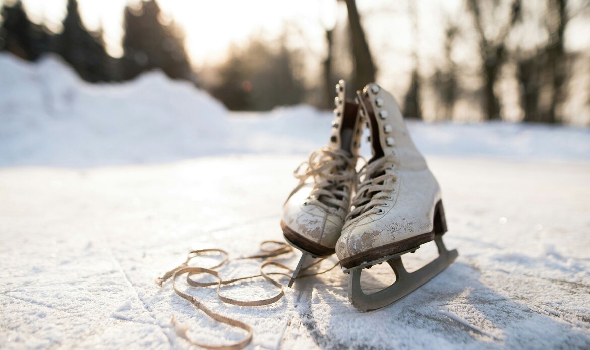 Ein Paar Eislaufschuhe steht auf einer glatten Eisfläche im Winterlicht, im Hintergrund sind Schnee und Bäume zu sehen.