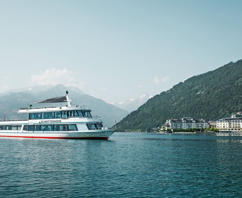 Ein Ausflugsschiff fährt über den Zeller See vor Bergen und einer Uferbebauung.