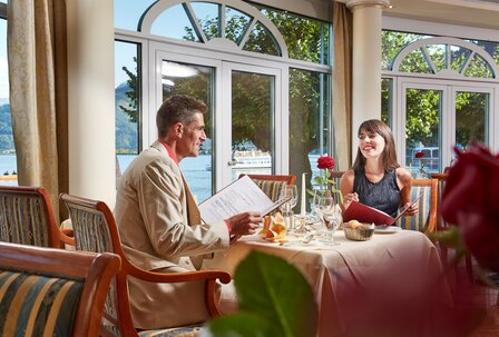 Hotelrestaurant mit Seeblick im GRAND HOTEL Ein Paar sitzt im Hotelrestaurant am Tisch mit Speisekarten und Blick auf den See durch große Fenster.