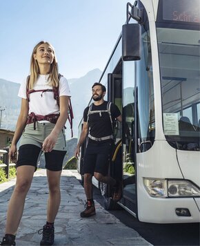 Eine Frau und ein Mann steigen neben einem Bus mit Wanderausrüstung aus, im Hintergrund sind Berge zu sehen.