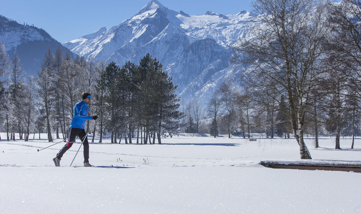 Ein Langläufer zieht auf einer verschneiten Fläche seine Spur vor einer Bergkulisse.