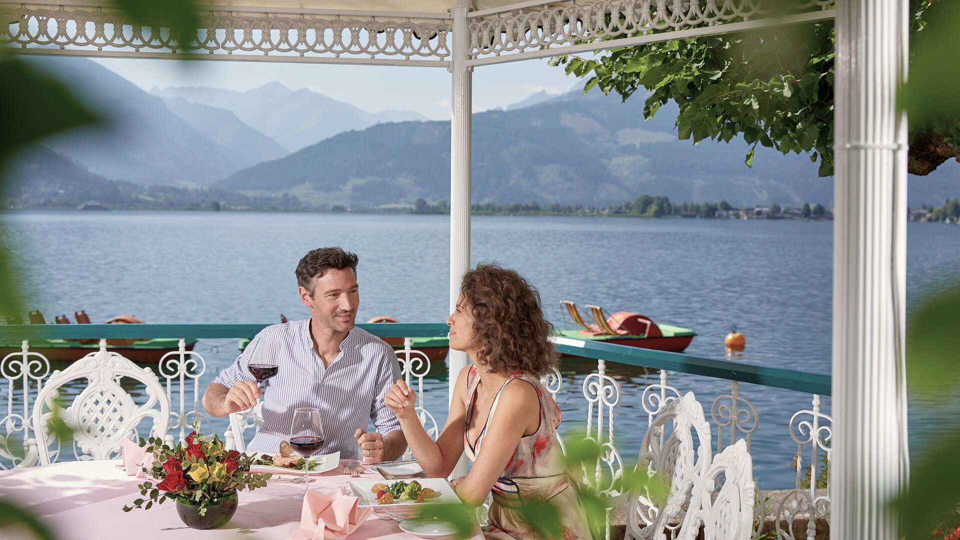 Ein Paar sitzt auf einer Restaurantterrasse am Zeller See und stößt mit Getränken an.