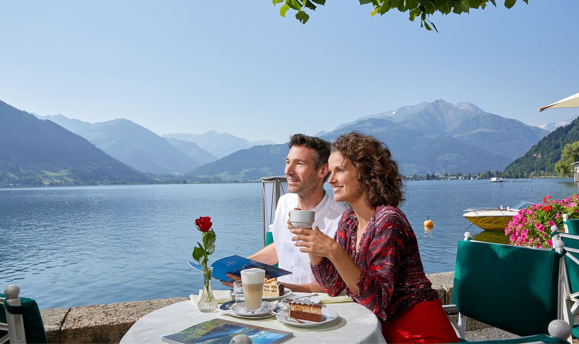 Ein Paar sitzt an einem Tisch am See und genießt Kaffee, Kuchen und Snacks mit Blick auf den Zeller See.