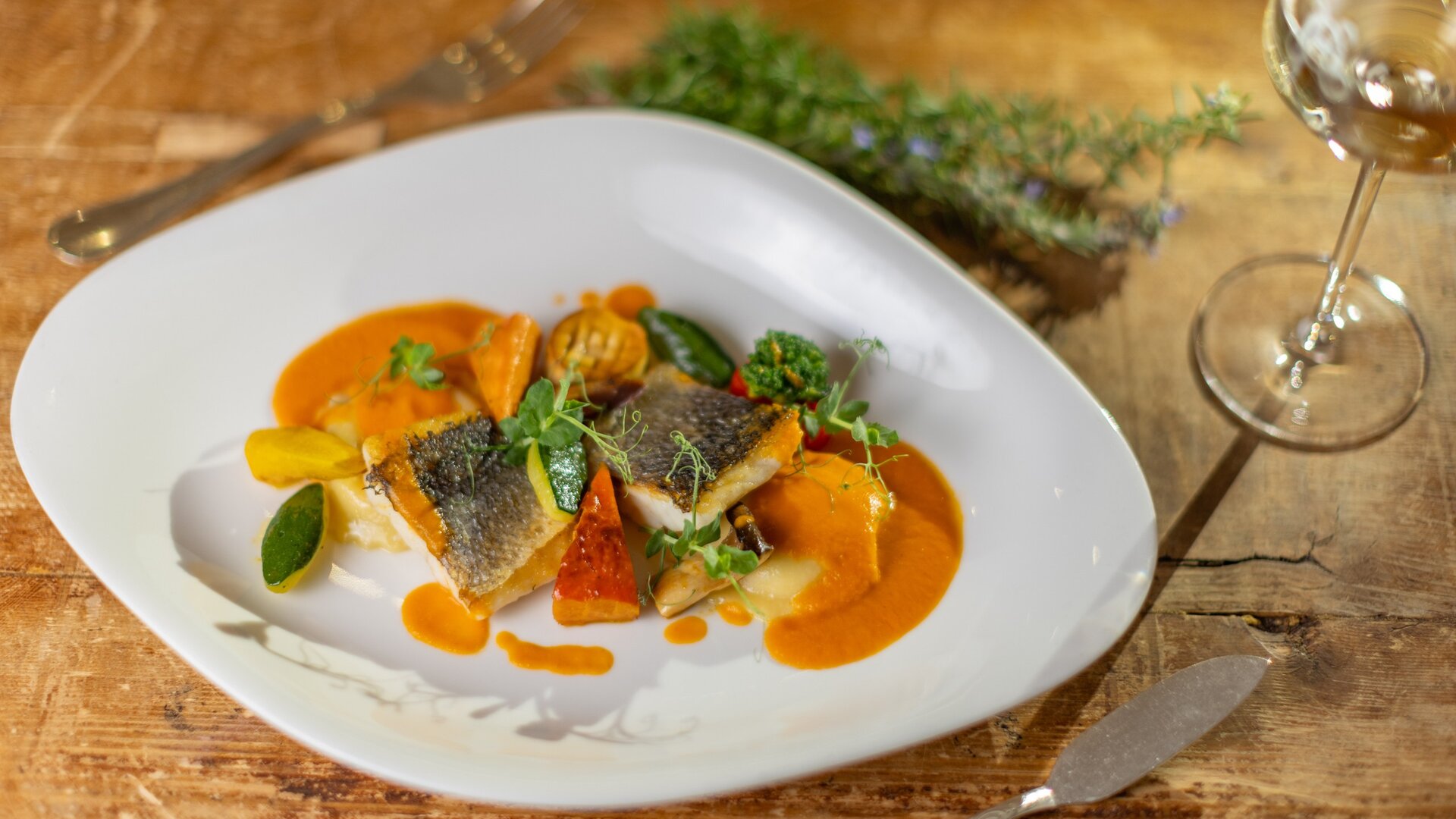Ein angerichtetes Fischgericht mit Gemüse und cremiger Sauce liegt auf einem weißen Teller auf einem Holztisch.