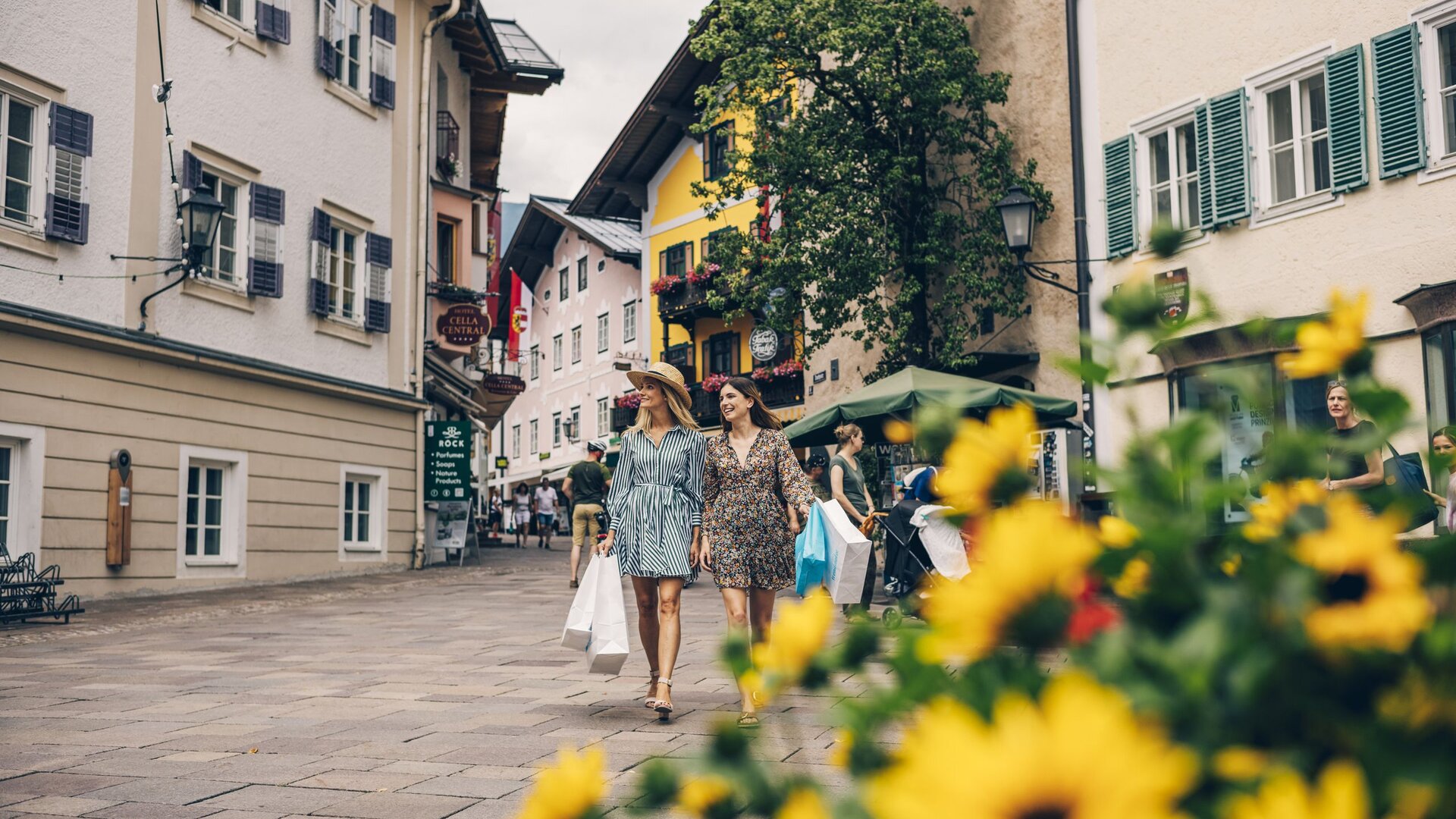Zwei Frauen spazieren durch eine Altstadtgasse in Zell am See, im Vordergrund blühen gelbe Blumen.