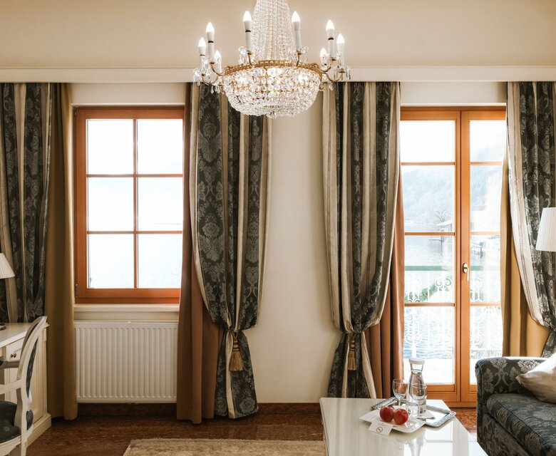 Ein elegantes Hotelzimmer mit Schreibtisch, Sofa, Kronleuchter und Balkonfenstern mit Blick nach draußen.