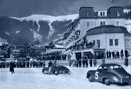 Ein historisches Schwarzweißfoto zeigt das GRAND HOTEL Zell am See mit Gästen und zwei Rennwagen vor dem verschneiten Gebäude.