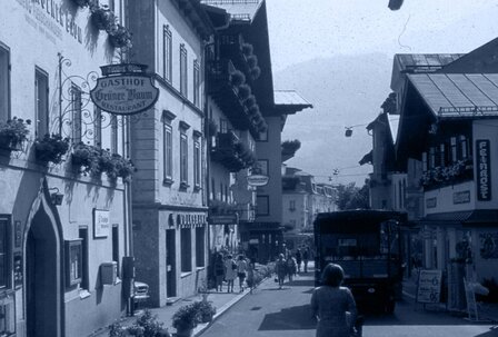 Eine historische Straße mit Gasthof-Schildern, Fußgängern und Fahrzeugen zwischen dicht stehenden Häusern.