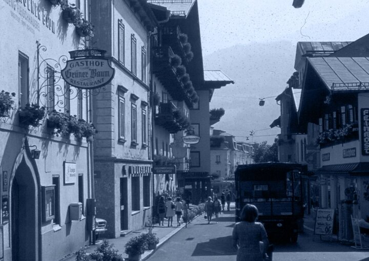 Eine historische Straße mit Gasthof-Schildern, Fußgängern und Fahrzeugen zwischen dicht stehenden Häusern.