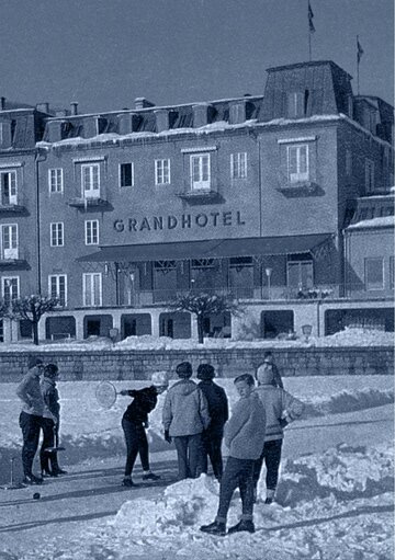 Mehrere Menschen stehen auf einer verschneiten Eisfläche vor dem GRANDHOTEL Zell am See.