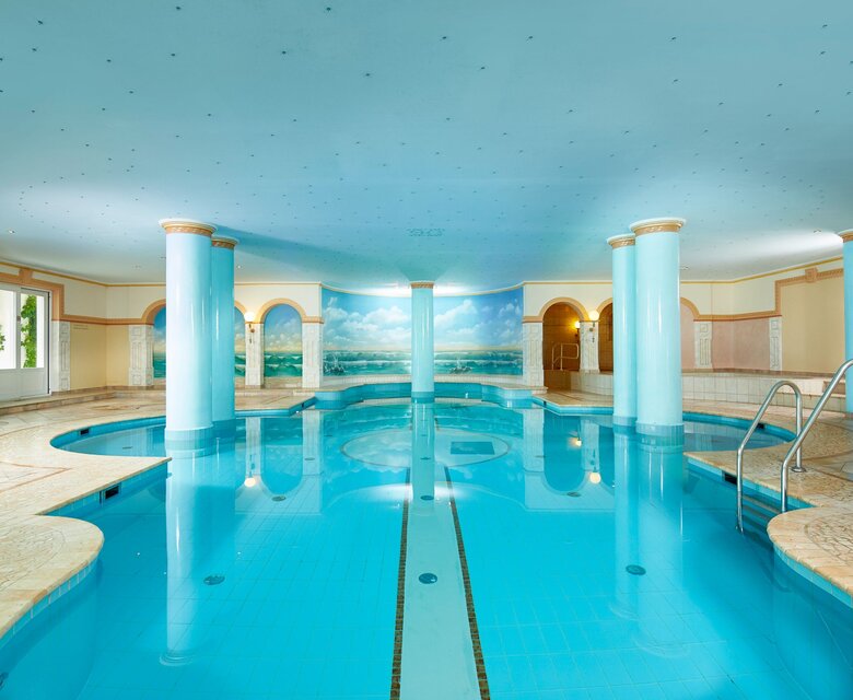 Der Innenpool mit türkisblauem Wasser liegt in einem hellen Wellnessbereich mit Säulen und Treppe.