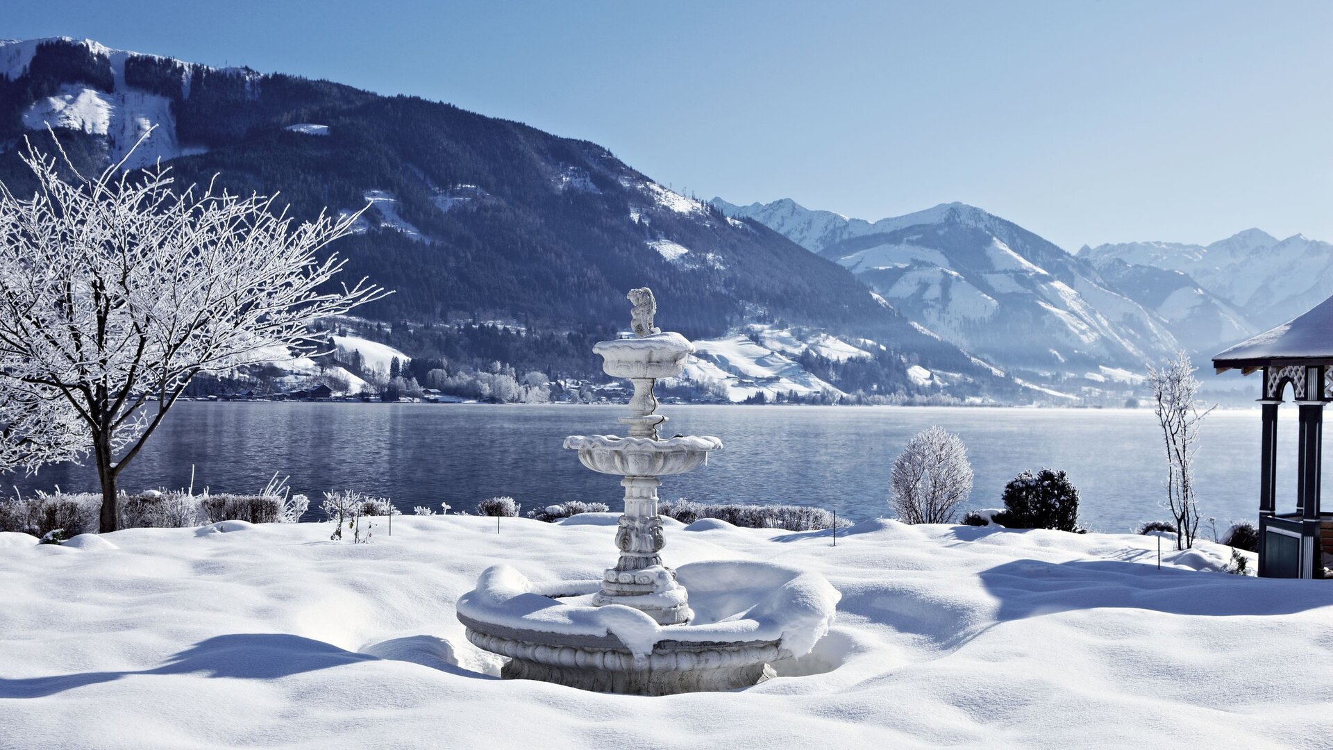 A snow-covered garden with a fountain stands by Lake Zell, with mountains in the winter background.