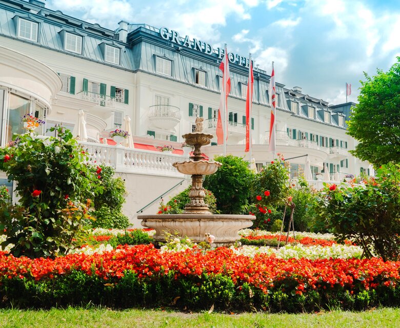 Ein gepflegter Hotelgarten mit Blumenbeeten und Springbrunnen vor dem GRAND HOTEL Zell am See.