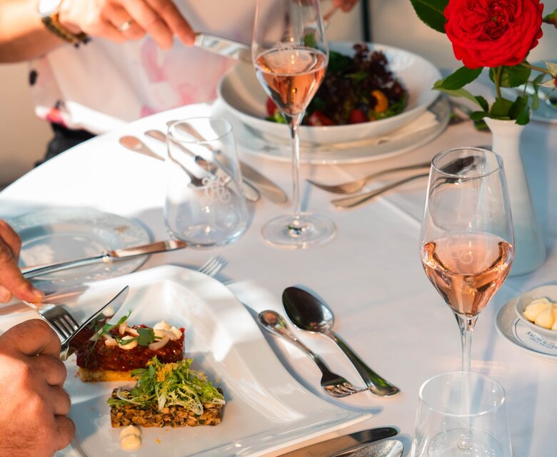 Ein festlich gedeckter Tisch im Hotelrestaurant mit zwei Gästen, Gläsern, Besteck und einem roten Blumenarrangement.
