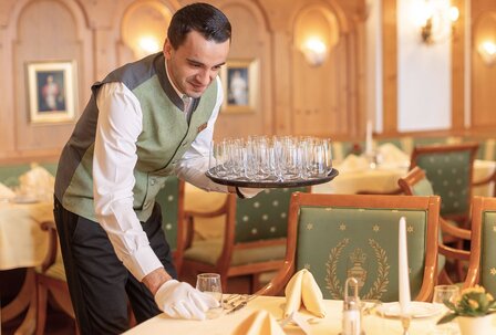 Servicemitarbeiter im Hotelrestaurant im GRAND HOTEL Zell am See Ein Servicemitarbeiter deckt im eleganten Hotelrestaurant einen Tisch und trägt Gläser auf einem Tablett.