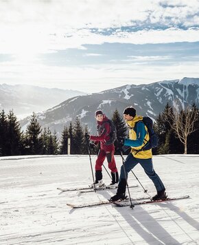 Zwei Langläufer stehen auf einer verschneiten Piste vor Bergen und winterlicher Landschaft bei Zell am See.