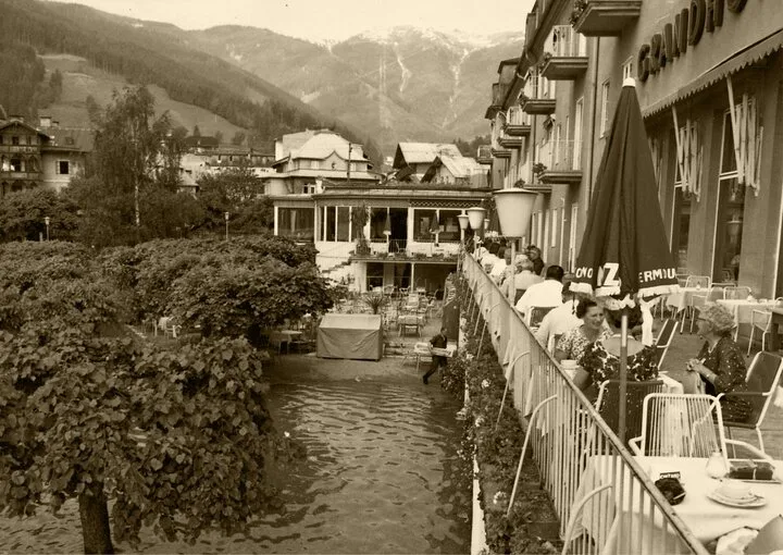 Eine historische Hotelterrasse mit Tischen und Stühlen am Wasser, im Hintergrund Berge und Gebäude.