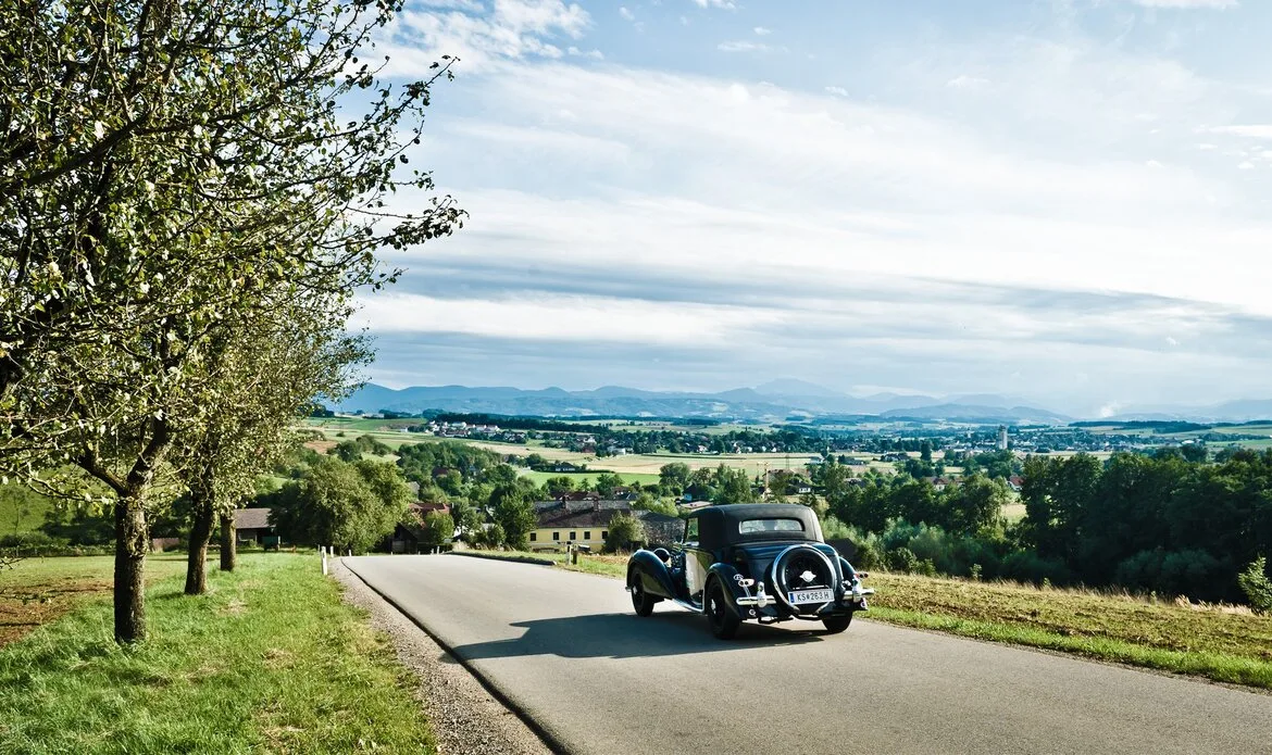 Ein Oldtimer fährt auf einer kurvigen Landstraße mit weitem Blick über grüne Hügel und das Tal.