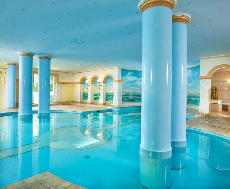 Ein heller Innenpool mit türkisfarbenem Wasser, hohen Säulen und Blick auf den See im GRAND HOTEL Zell am See.