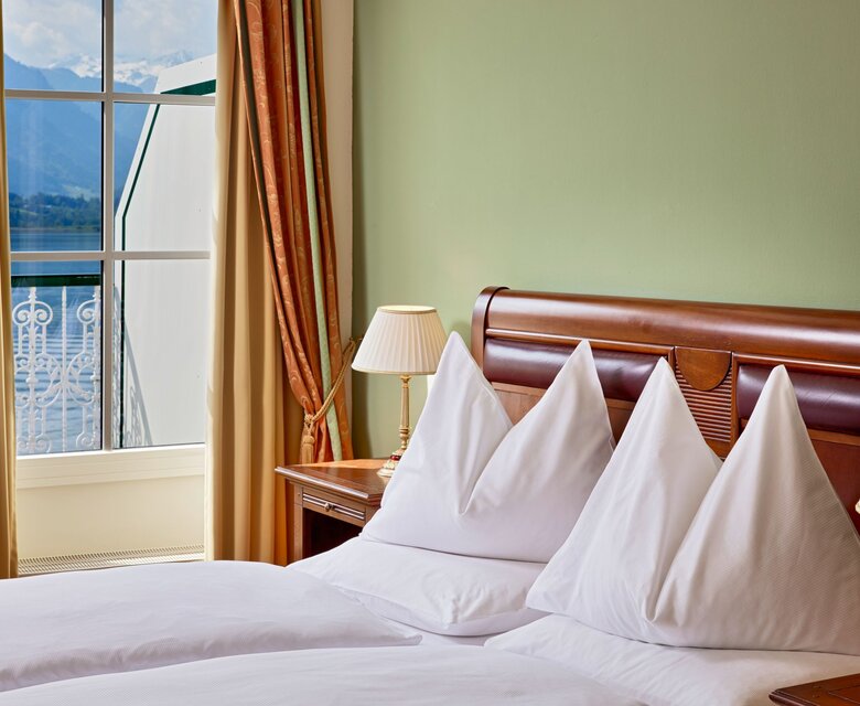 Ein Doppelbett mit weißen Kissen steht vor einem Fenster mit Blick auf den See und den Balkon.