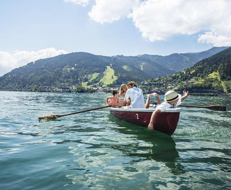 Mehrere Personen fahren in einem Ruderboot auf dem Zeller See, im Hintergrund liegen Berge und grüne Hänge.