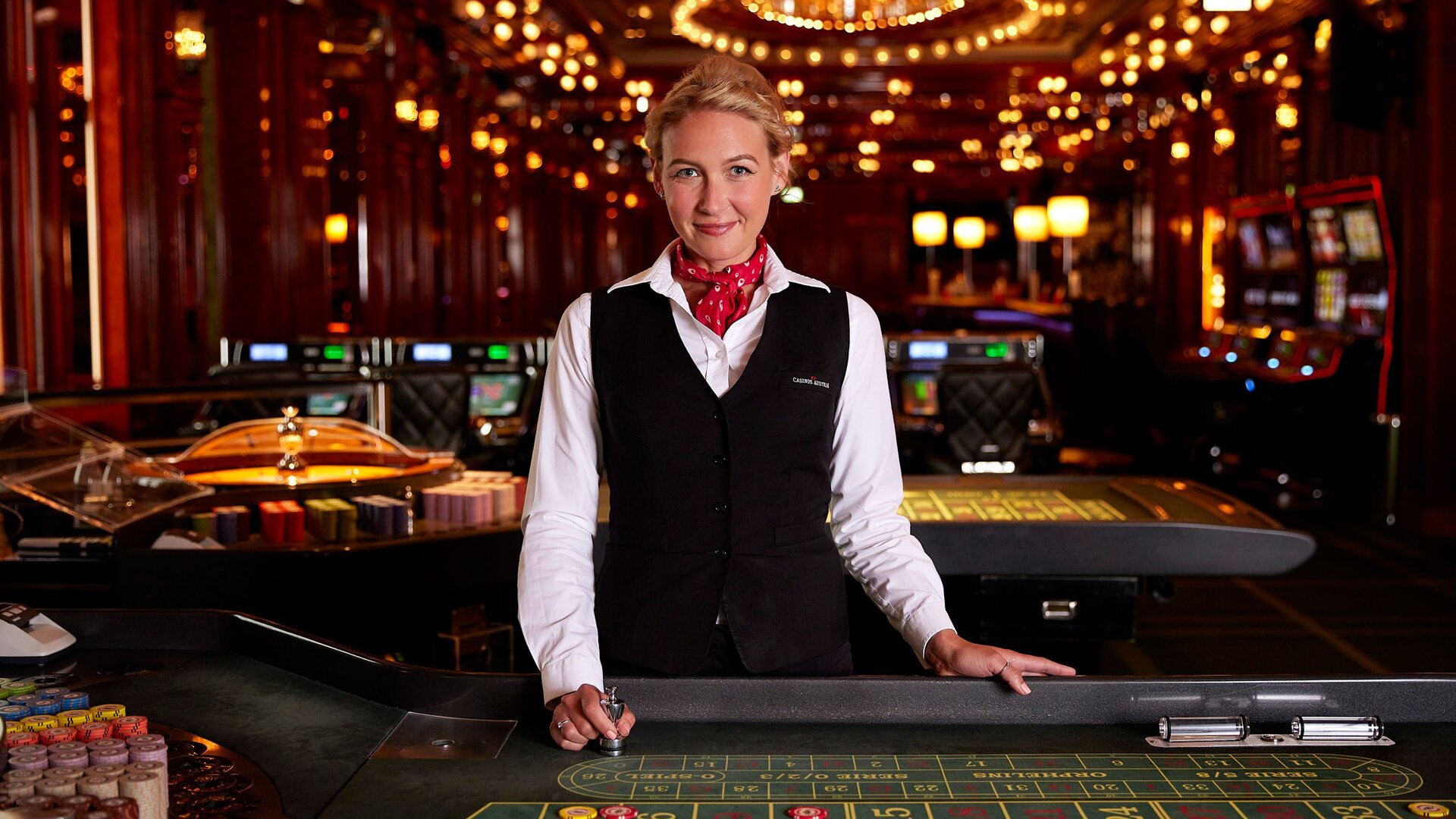 A staff member stands at a roulette table in Casino Zell am See, with gaming tables and slot machines behind her.