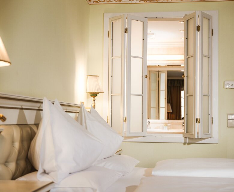 Ein helles Hotelzimmer mit aufgeschlagenem Bett, zwei Nachttischlampen und geöffneten Fenstern zum Nebenraum.